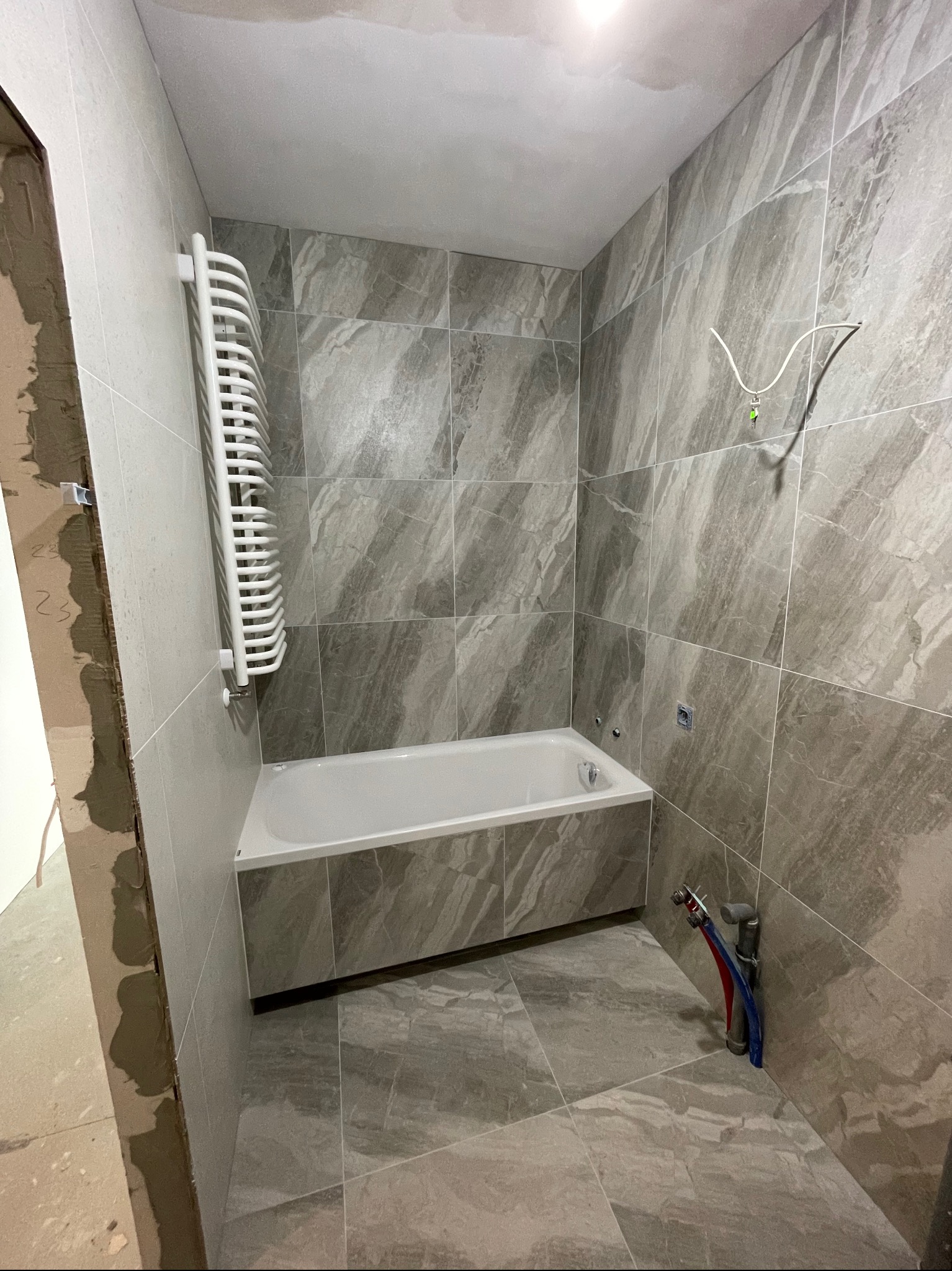 Łazienka w trakcie remontu: położone szare płytki imitujące marmur, biała wanna, grzejnik drabinkowy, widoczne wystające rury i przewody, surowy otwór drzwiowy.