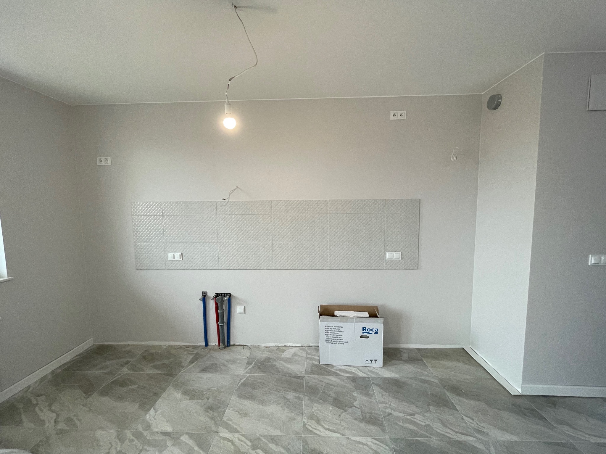 Wnętrze łazienki w trakcie remontu: ułożona podłoga z szarych płytek imitujących marmur, fragment ściany z zamontowaną dekoracyjną płytką ceramiczną z geometrycznym wzorem, widoczne rury instalacji...