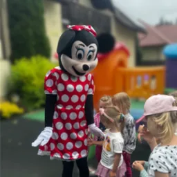 Osoba przebrana za postać z bajki w czerwonej sukience w białe grochy i żółtych butach, otoczona grupą dzieci na tle dmuchanego zamku.