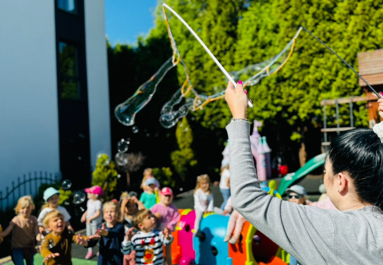 Kobieta puszcza duże bańki mydlane, wokół grupa roześmianych dzieci na tle zieleni i placu zabaw.