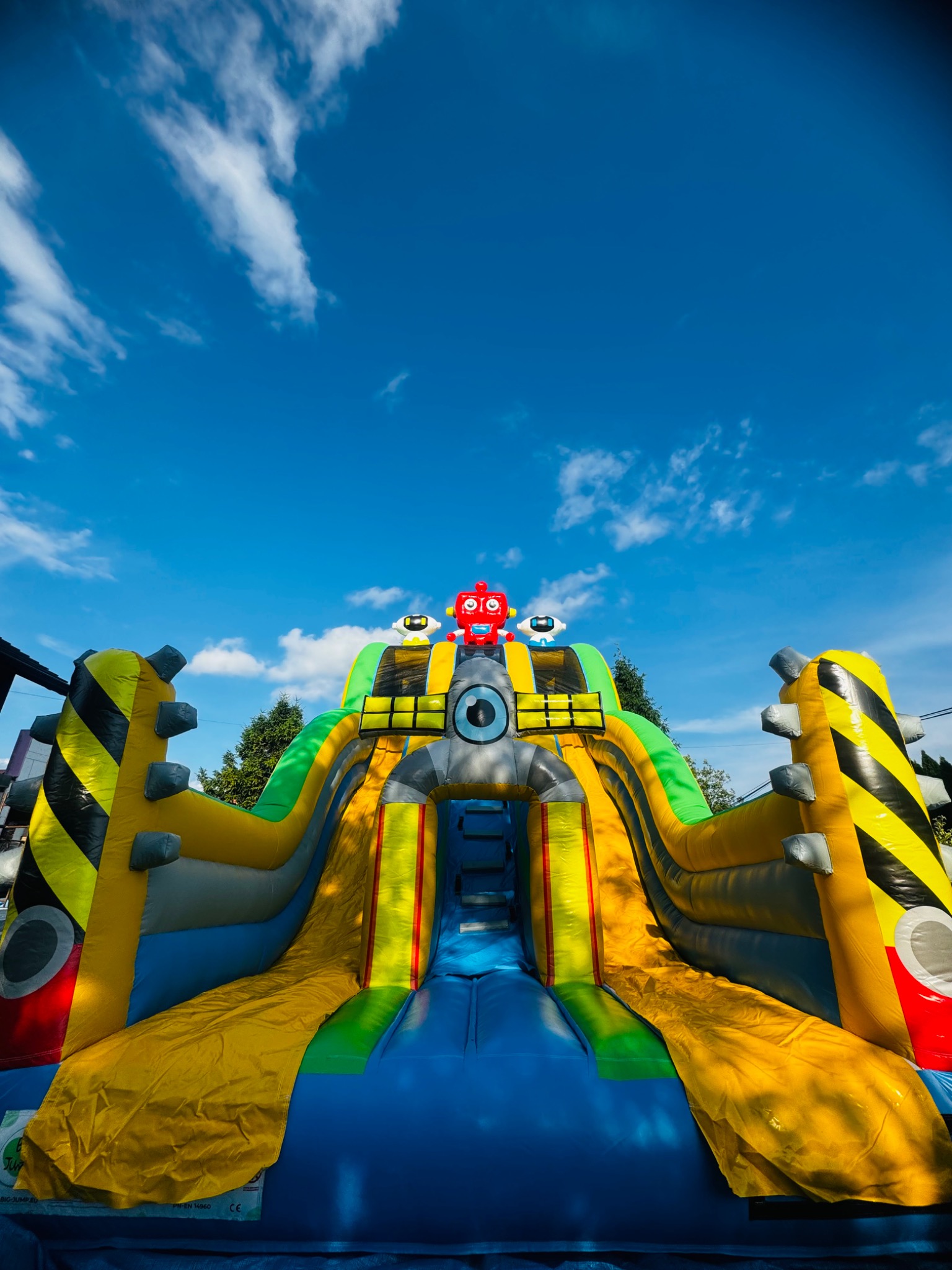 Kolorowa, dmuchana zjeżdżalnia w kształcie rakiety z robotami na szczycie, na tle błękitnego nieba z chmurami.