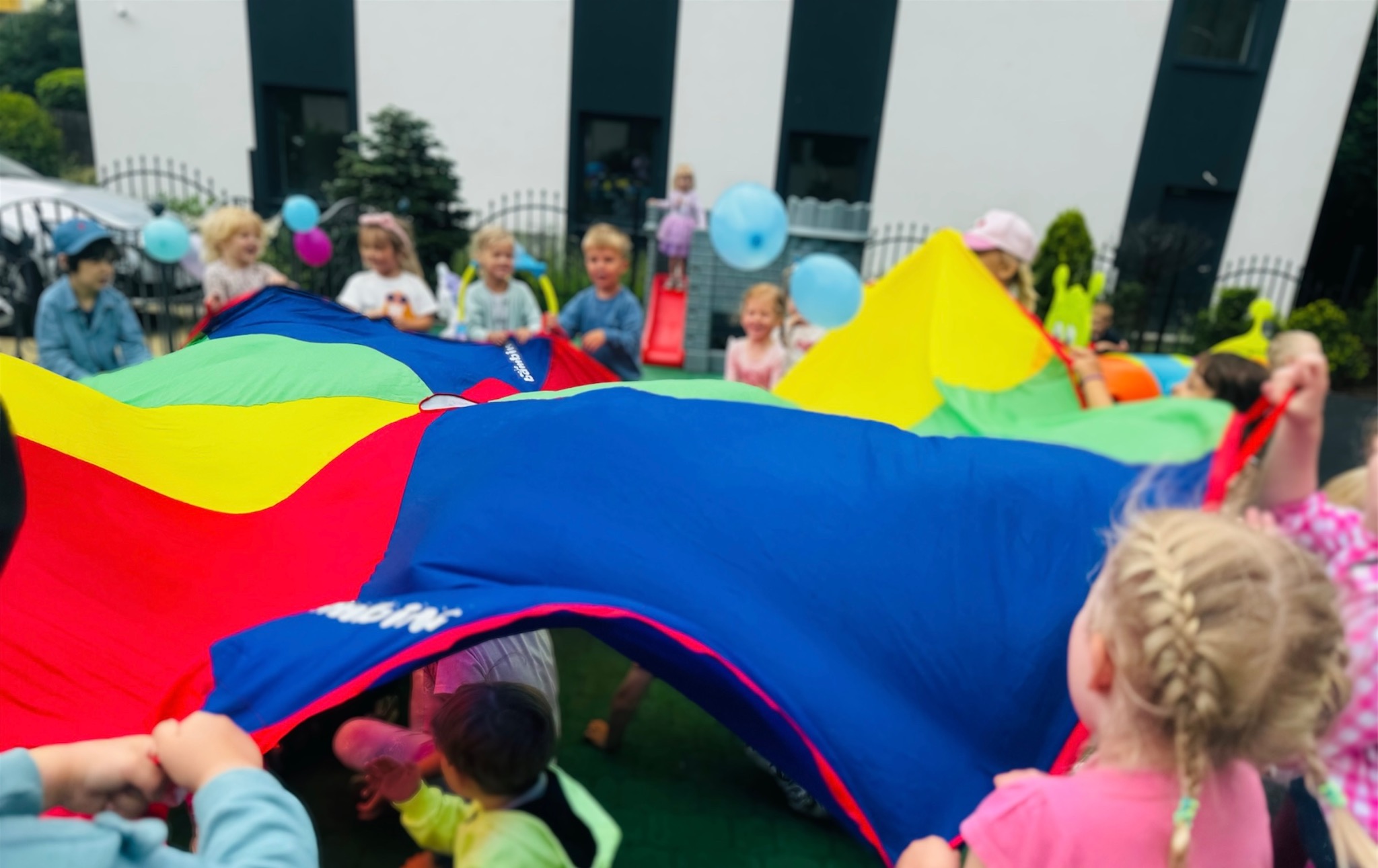 Grupa dzieci bawi się kolorową chustą animacyjną na świeżym powietrzu, w tle widoczny budynek z czarno-białymi pasami i plac zabaw.