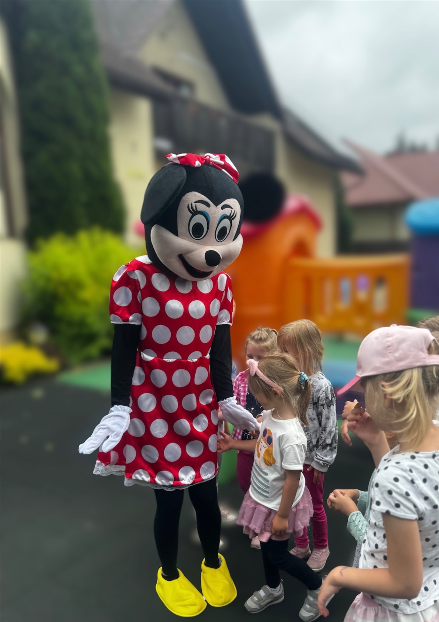 Osoba przebrana za postać z bajki w czerwonej sukience w białe grochy i żółtych butach, otoczona grupą dzieci na tle dmuchanego zamku.