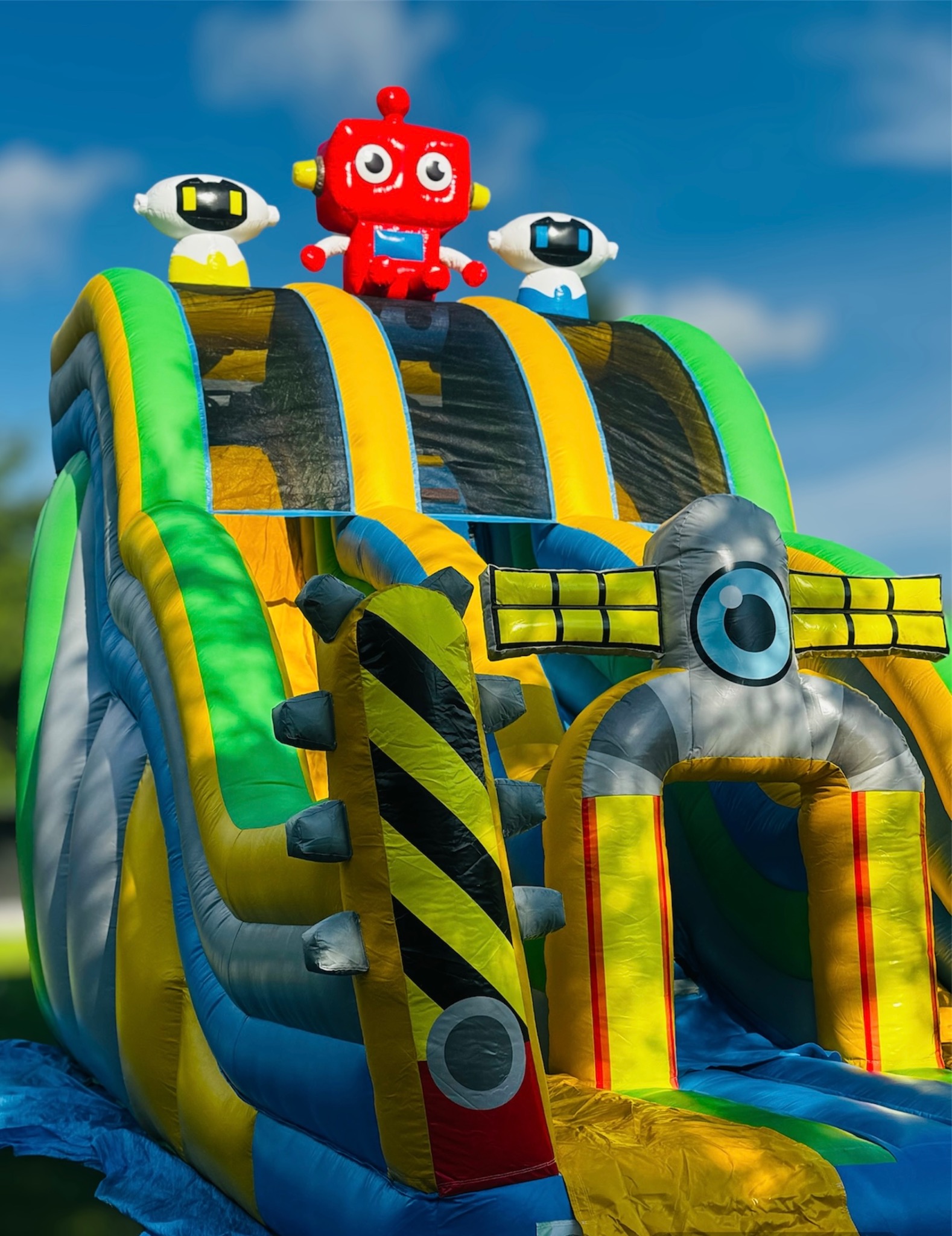Kolorowa dmuchana zjeżdżalnia z motywem robotów, w odcieniach zieleni, żółci i błękitu, z trzema robotami na szczycie: czerwonym pośrodku i dwoma białymi po bokach, na tle błękitnego nieba.