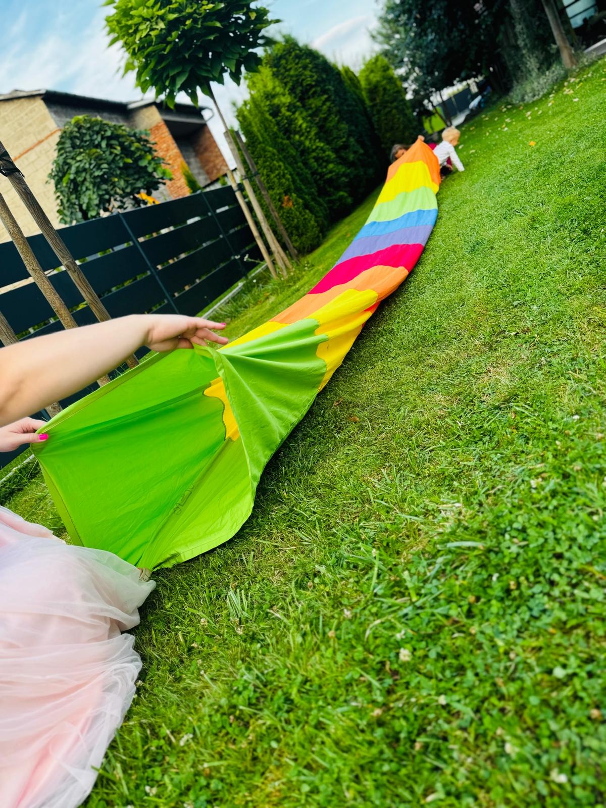 Dzieci bawią się na trawie w tunelu z kolorowej tkaniny trzymanej przez osobę dorosłą.