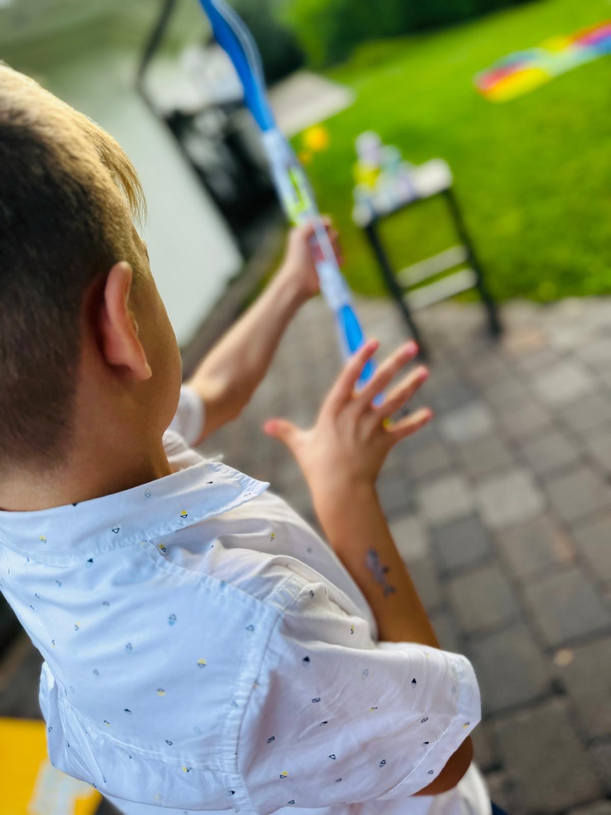 Chłopiec w białej koszuli z krótkim rękawem, z tatuażem rekina na ramieniu, bawi się niebieską zabawką na tle zielonego trawnika i rozmytego stołu piknikowego.