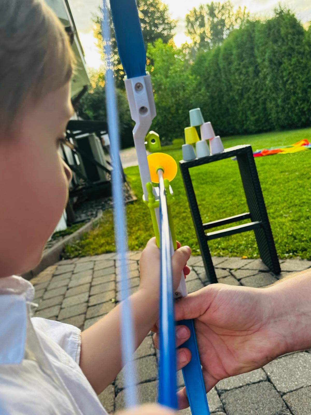 Chłopiec celuje z plastikowego łuku do piramidy z kubków na tle zielonego trawnika i żywopłotu.