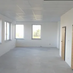 Puste, jasne pomieszczenie biurowe z szarą podłogą z płytek, białymi ścianami, trzema oknami i podwieszanym sufitem kasetonowym z widoczną kratką wentylacyjną.