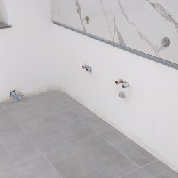 Filmar - Wnętrze kuchni w trakcie wykańczania: szare płytki podłogowe, białe ściany z fragmentem marmurowych płytek nad blatem i wyprowadzonymi instalacjami hydraulicznymi.