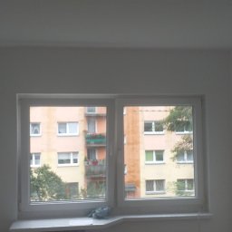 Filmar - Wnętrze pokoju z nowym, białym oknem PCV; widok na blok mieszkalny z zielonymi balkonami i drzewami za oknem; ściany w trakcie malowania na biało, widoczna listwa przypodłogowa oparta o ścianę.