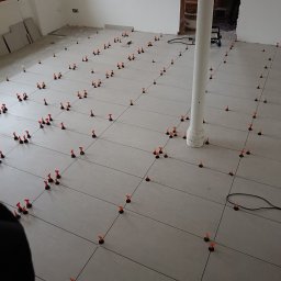 Filmar - Układanie szarych płytek podłogowych z użyciem systemu poziomowania, widoczny biały filar w pomieszczeniu w trakcie prac remontowych.