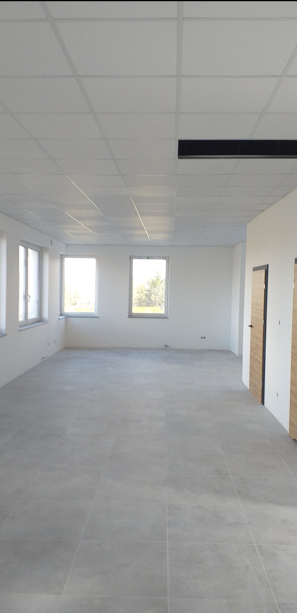 Puste, jasne pomieszczenie biurowe z szarą podłogą z płytek, białymi ścianami, trzema oknami i podwieszanym sufitem kasetonowym z widoczną kratką wentylacyjną.