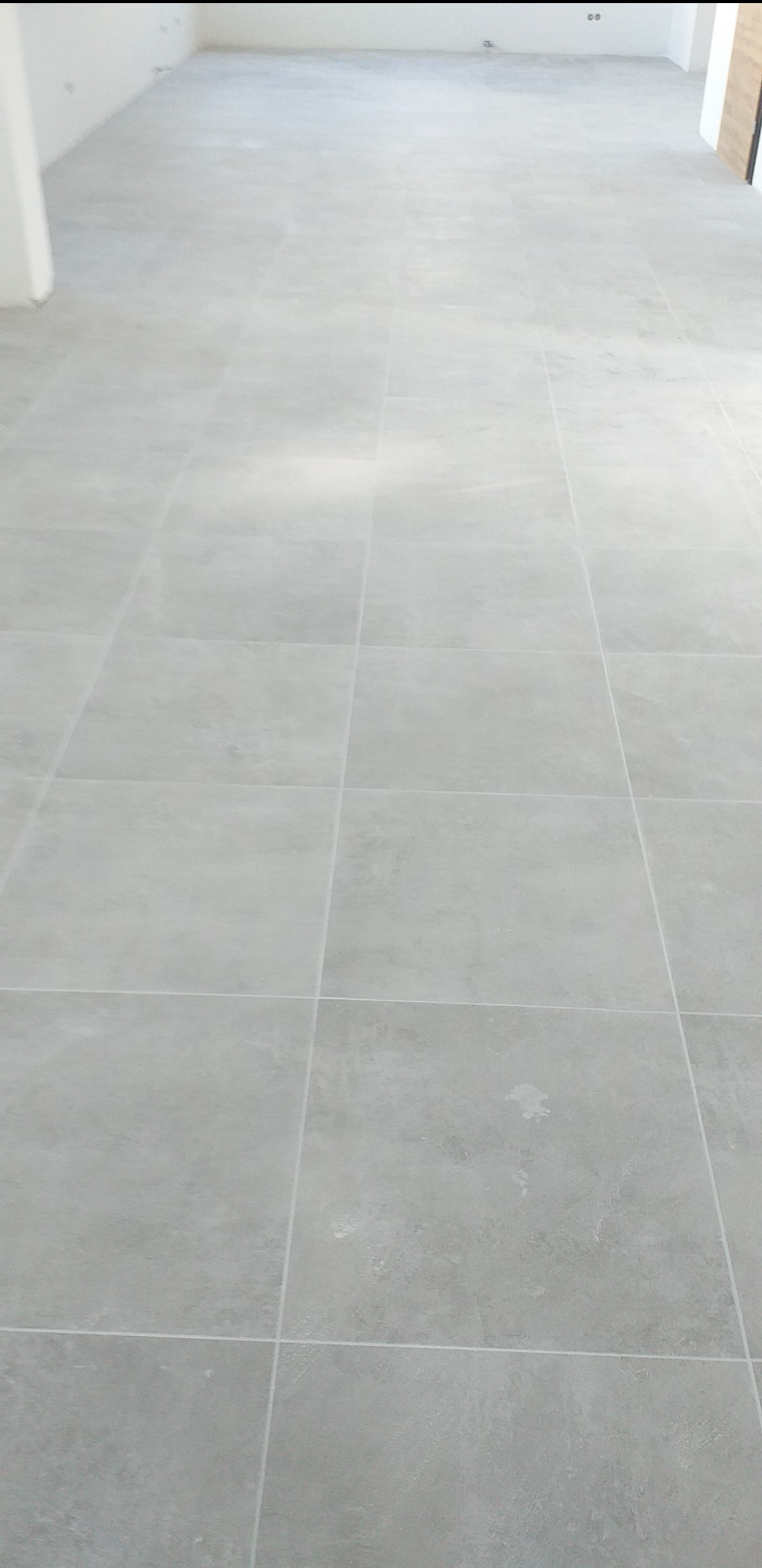 Jasnoszara, świeżo ułożona podłoga z płytek ceramicznych w pustym pomieszczeniu, widoczne białe fugi, na niektórych płytkach widoczne drobne zabrudzenia.