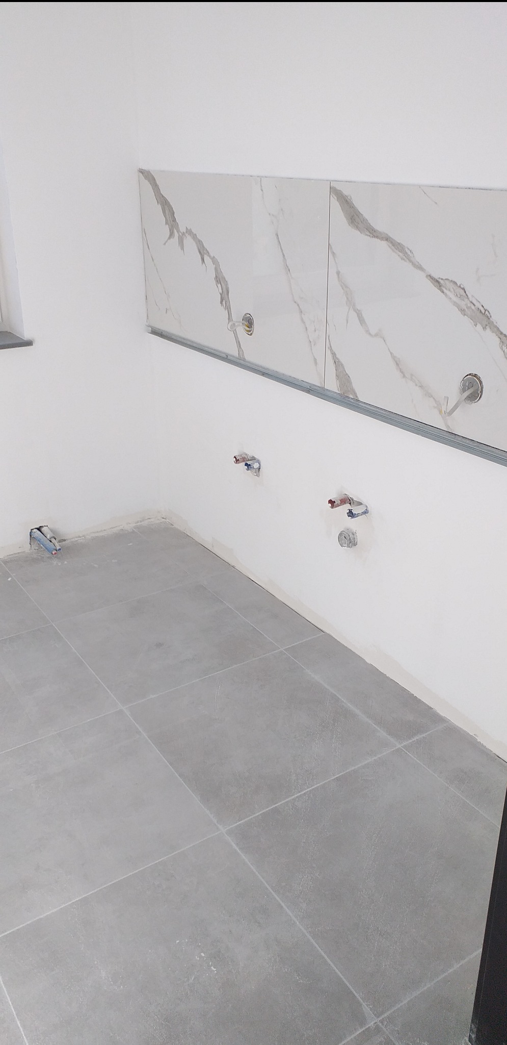 Wnętrze kuchni w trakcie wykańczania: szare płytki podłogowe, białe ściany z fragmentem marmurowych płytek nad blatem i wyprowadzonymi instalacjami hydraulicznymi.