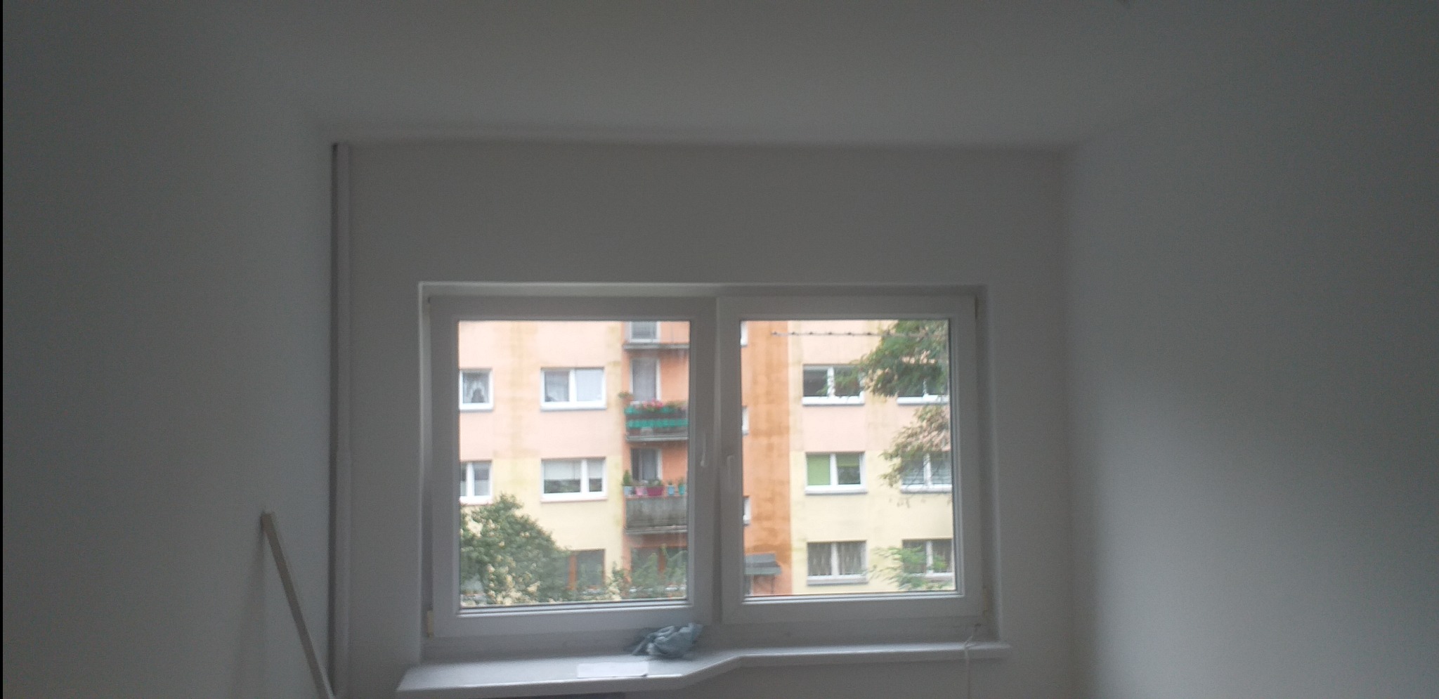 Wnętrze pokoju z nowym, białym oknem PCV; widok na blok mieszkalny z zielonymi balkonami i drzewami za oknem; ściany w trakcie malowania na biało, widoczna listwa przypodłogowa oparta o ścianę.