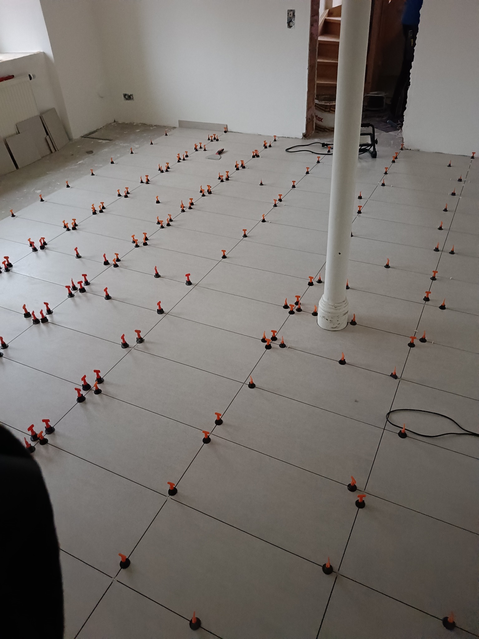 Układanie szarych płytek podłogowych z użyciem systemu poziomowania, widoczny biały filar w pomieszczeniu w trakcie prac remontowych.