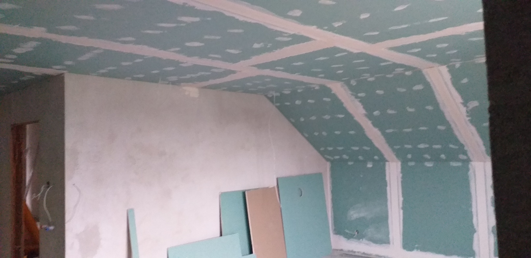 Poddasze w trakcie wykańczania płytami gipsowo-kartonowymi; widoczne łączenia płyt wypełnione masą szpachlową, niezagruntowana ściana szczytowa i nieużyte płyty opierające się o ścianę.