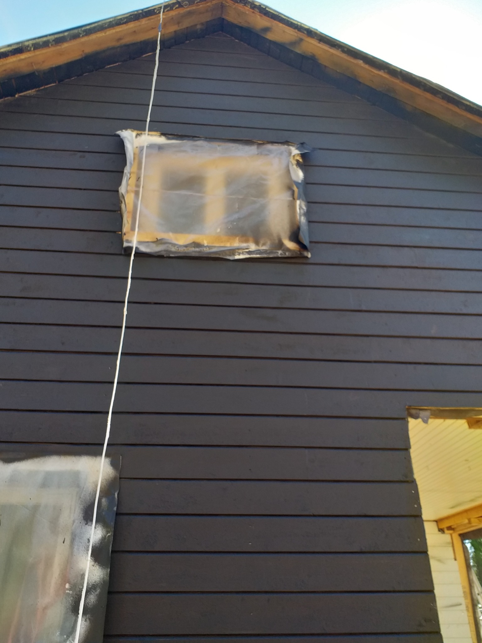 Fragment ściany zewnętrznej domu obitego ciemnymi deskami, z oknem zabezpieczonym folią malarską i fragmentem niezabezpieczonego otworu okiennego, widoczne ślady po malowaniu.