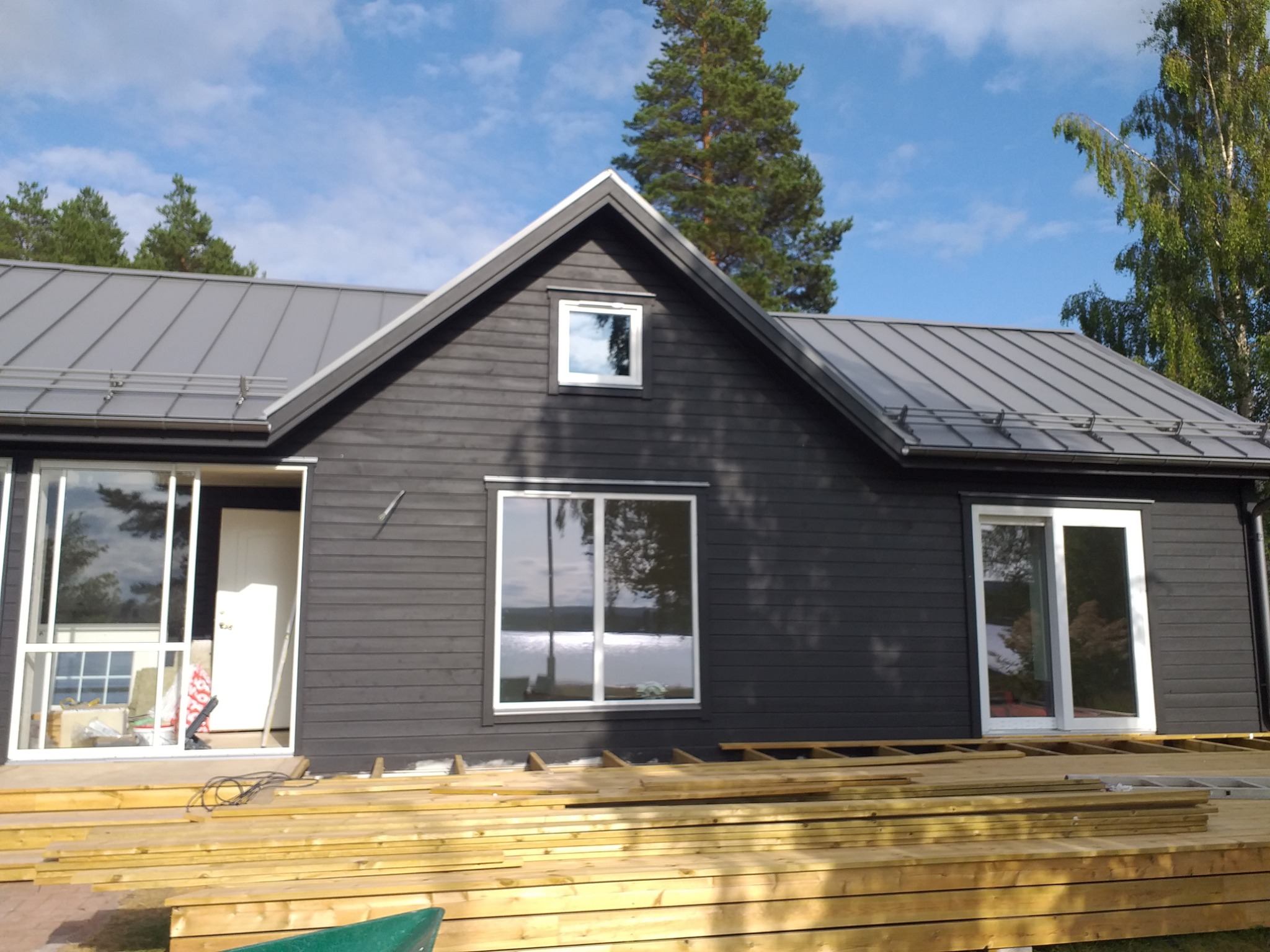 Dom z ciemną drewnianą elewacją i szarym dachem, w trakcie budowy tarasu z surowego drewna, widok na okna z odbiciem jeziora i nieba.