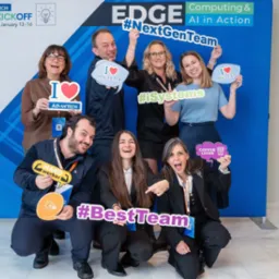 Grupa uśmiechniętych osób pozuje na tle firmowej ścianki reklamowej z hasłami #NextGenTeam, #iSystems, #BestTeam i rekwizytami 'I love my team', 'Hard worker', 'Coffee lover' podczas wydarzenia...