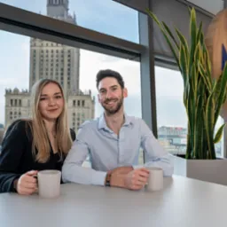 Dwoje uśmiechniętych pracowników biurowych siedzi przy stole z kubkami, z widokiem na Pałac Kultury i Nauki w Warszawie, obok logo firmy MARS i doniczki z rośliną.