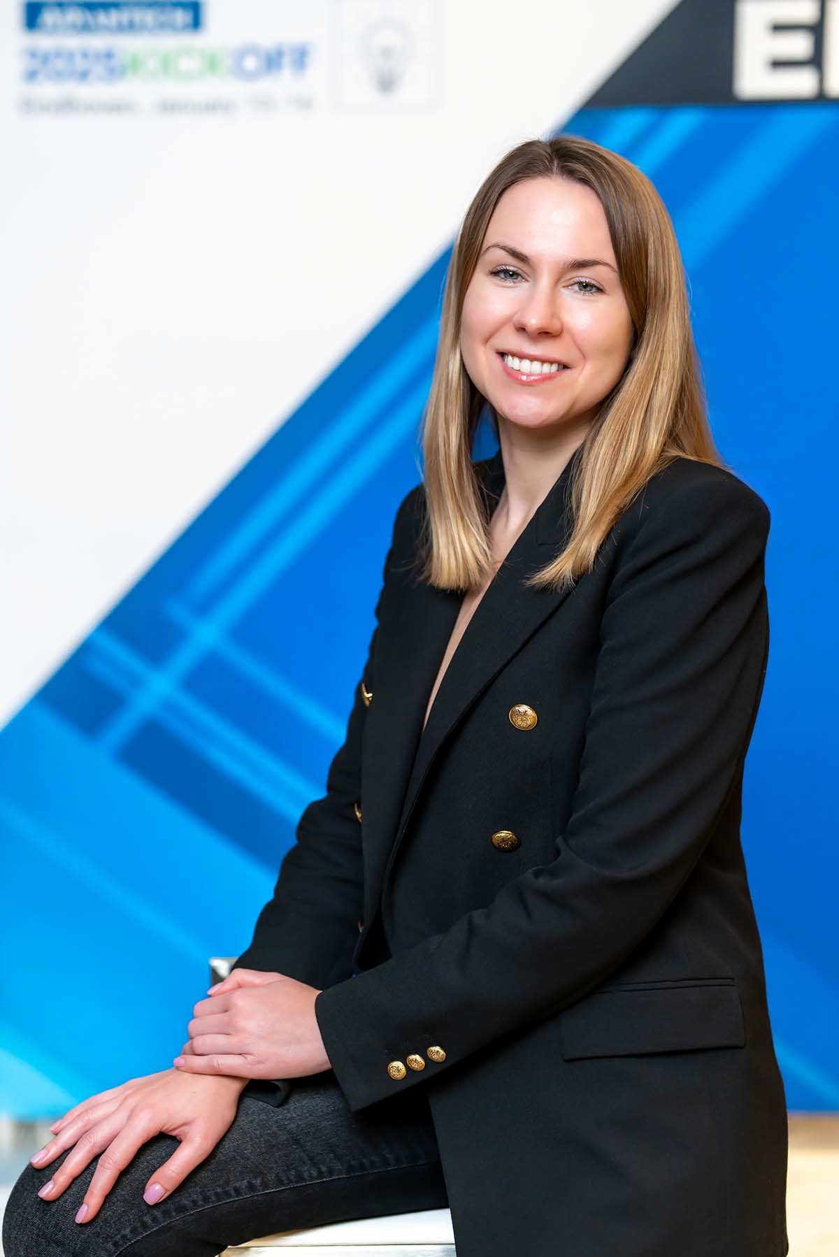 Portret uśmiechniętej kobiety w czarnej marynarce z ozdobnymi guzikami na tle białej ściany z niebieskimi akcentami.