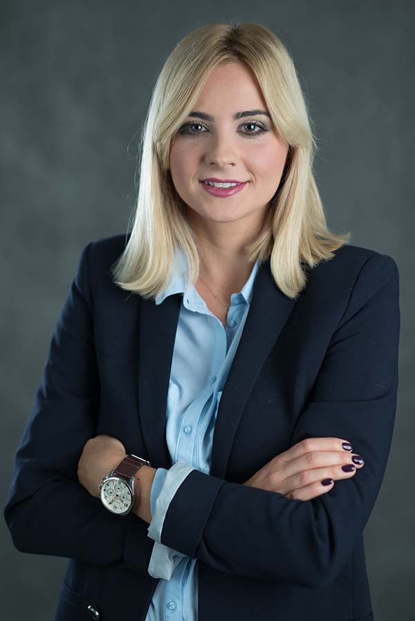 Portret uśmiechniętej blondynki w granatowej marynarce i błękitnej koszuli, z zegarkiem na ręku, na szarym tle.