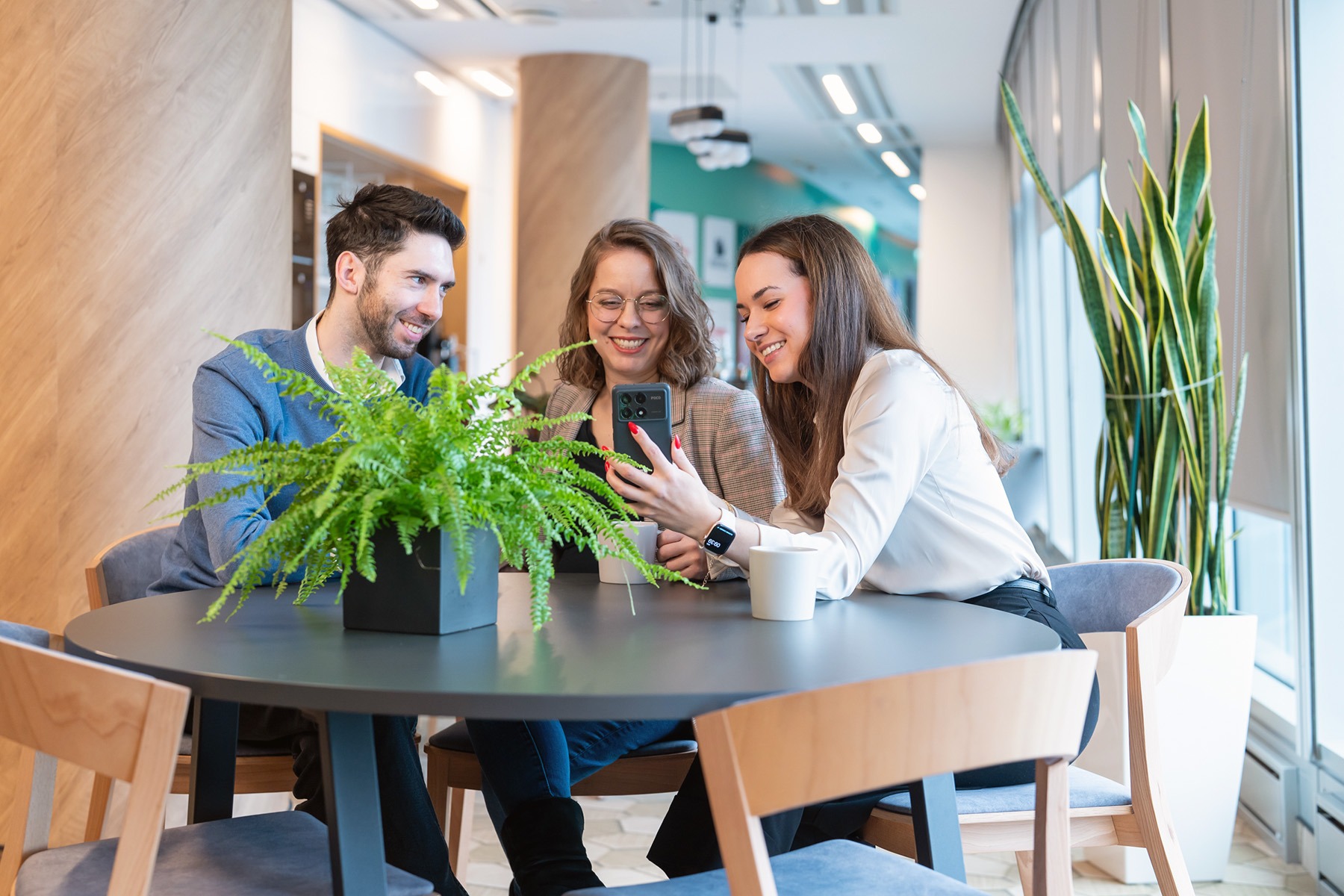Trzy osoby w biurze, siedzące przy okrągłym stole z paprotką, przeglądają zawartość smartfona, uśmiechając się.
