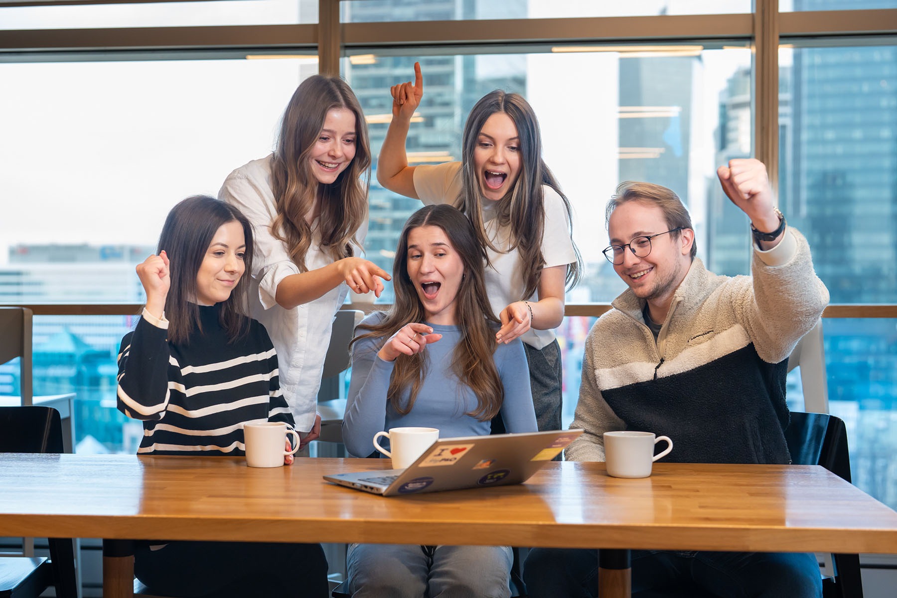 Zespół pięciu osób, w tym jedna siedząca przed laptopem z naklejkami, świętuje sukces przy drewnianym stole z kubkami, na tle okien z widokiem na miasto.