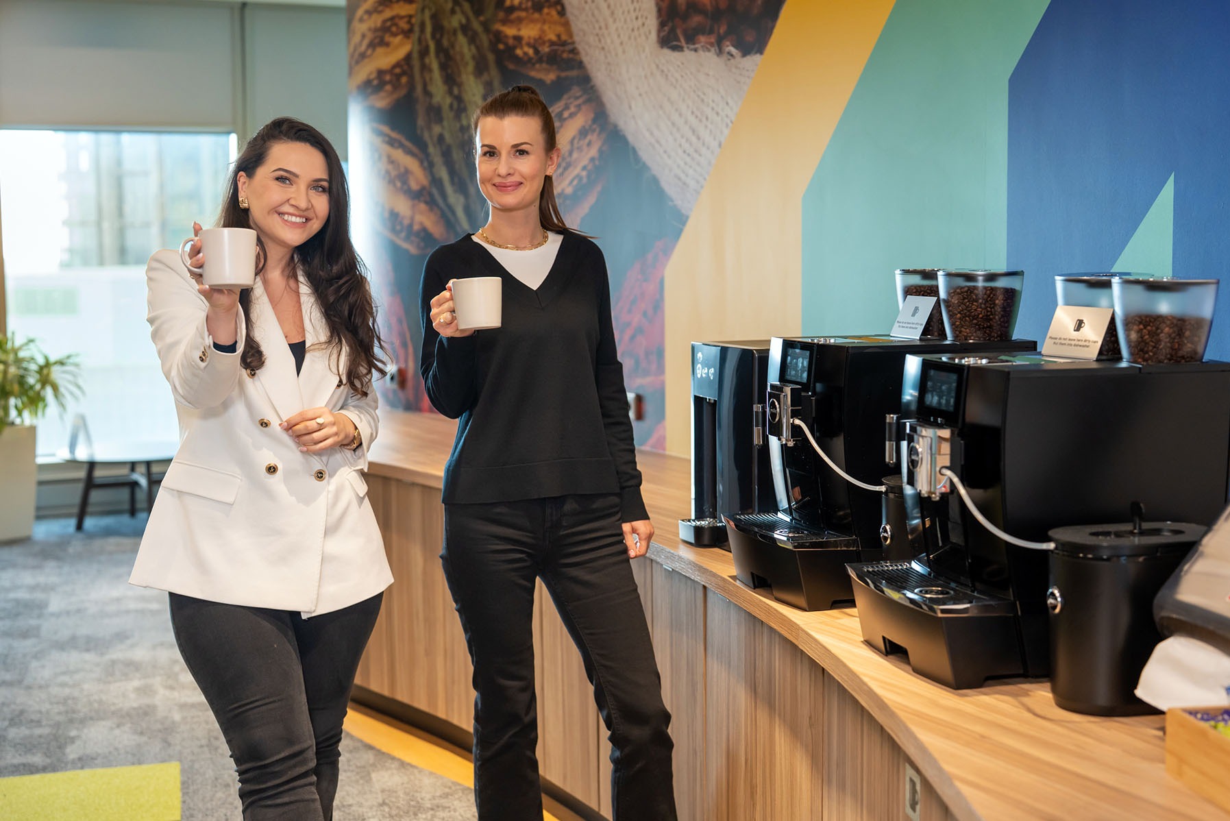 Dwie kobiety w biurze, stojące obok ekspresów do kawy, trzymają białe kubki. W tle grafika z motywem kawy i ziaren.
