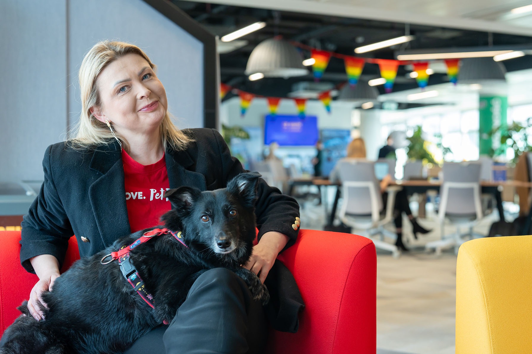 Kobieta w czarnej marynarce i czerwonej koszulce z psem rasy mieszanej siedząca na czerwonym fotelu w nowoczesnym biurze z dekoracjami w barwach tęczy.