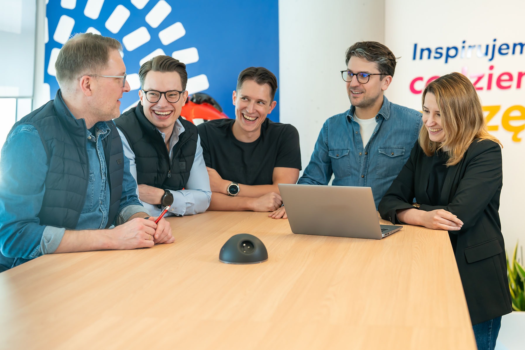 Zespół pięciu uśmiechniętych osób w Warszawie siedzi przy jasnym stole z laptopem, dyskutując i patrząc w lewo, z dekoracyjnym napisem na ścianie w tle.
