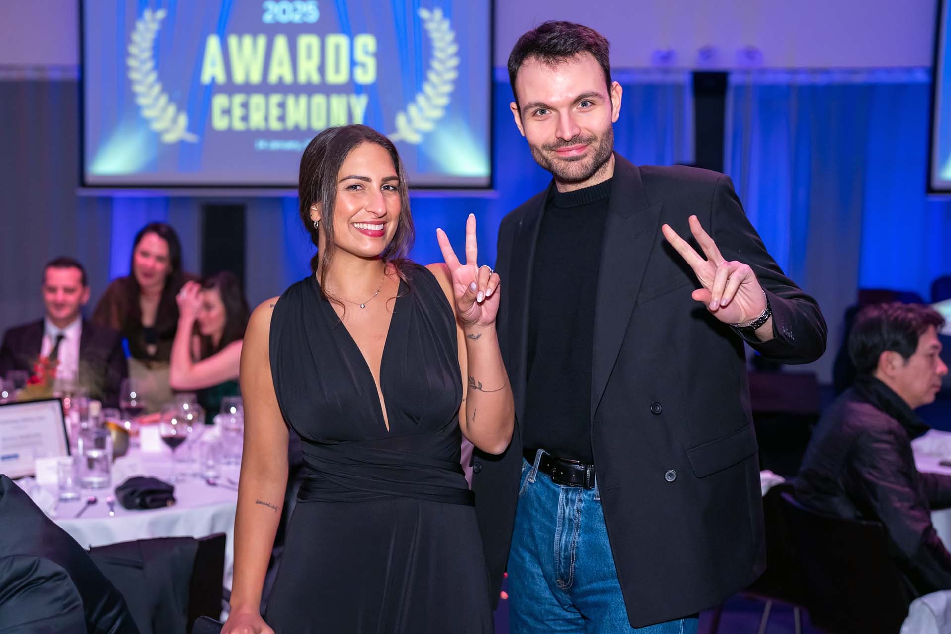 Dwie osoby, kobieta w czarnej sukience i mężczyzna w czarnej marynarce i jeansach, pozują na tle sali bankietowej z napisem '2025 Awards Ceremony' na ekranie, oboje pokazują znak 'V'.