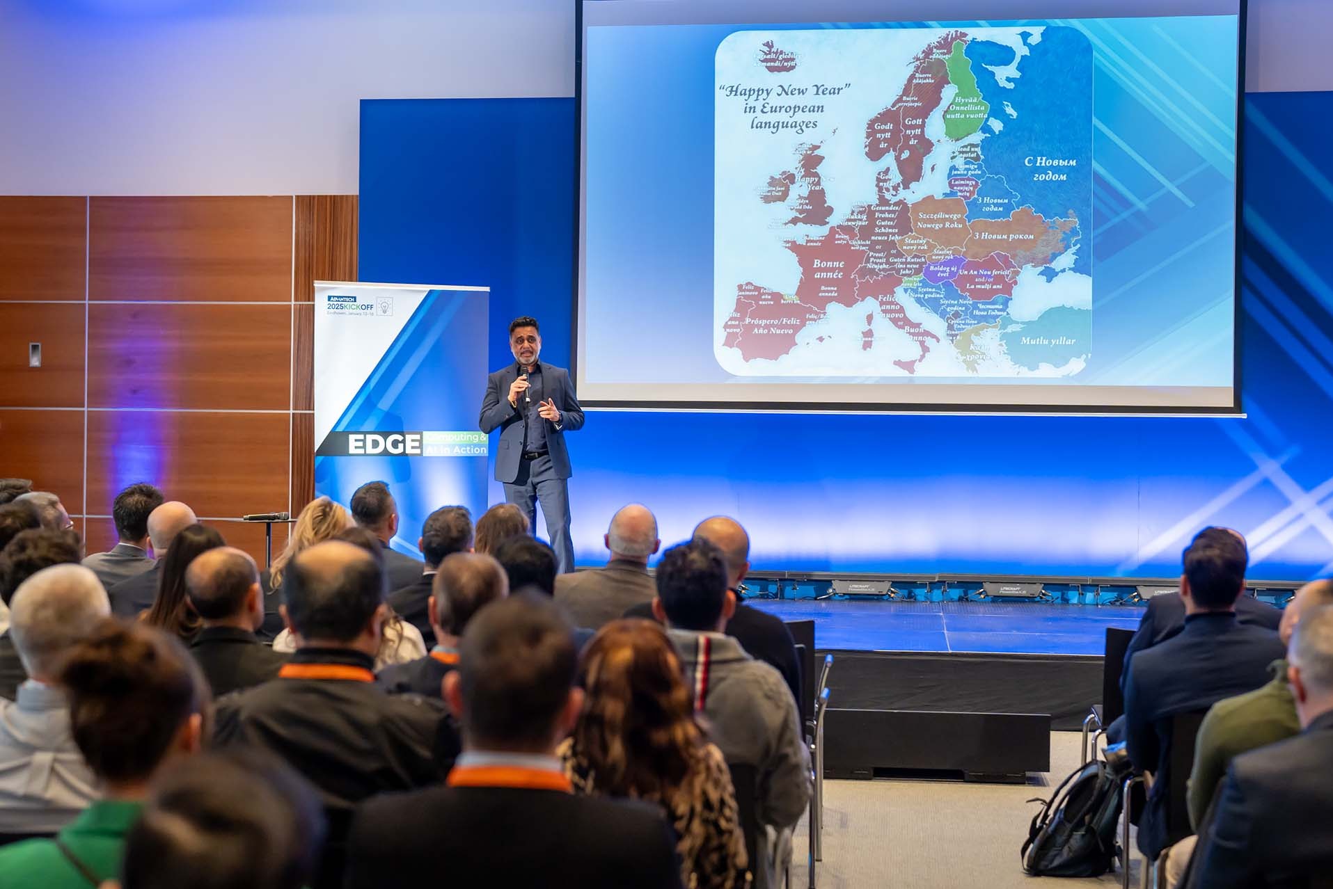 Prelegent w garniturze przemawia do publiczności podczas prezentacji z mapą Europy i życzeniami noworocznymi w różnych językach wyświetlanymi na ekranie.