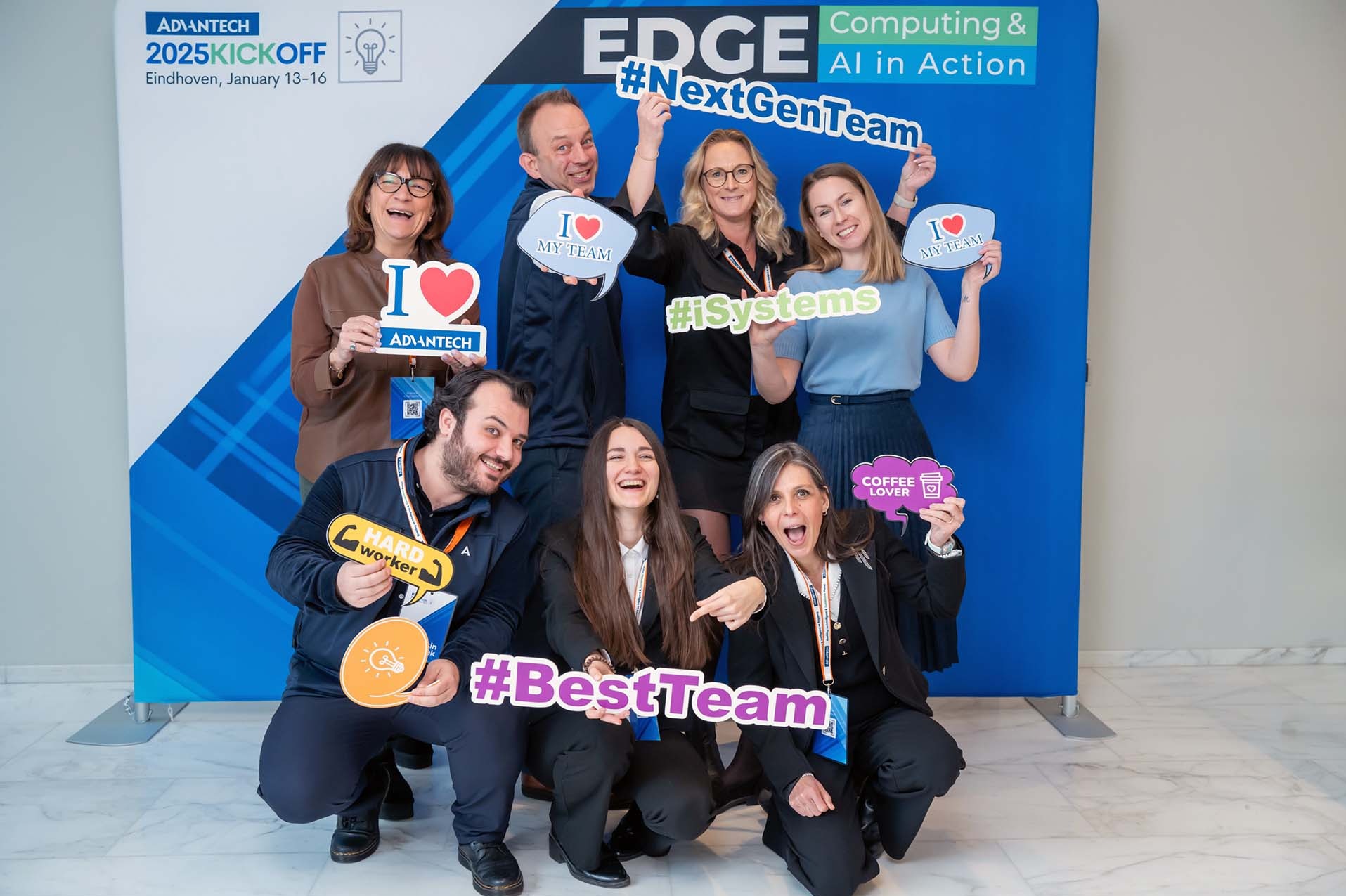 Grupa uśmiechniętych osób pozuje na tle firmowej ścianki reklamowej z hasłami #NextGenTeam, #iSystems, #BestTeam i rekwizytami 'I love my team', 'Hard worker', 'Coffee lover' podczas wydarzenia...