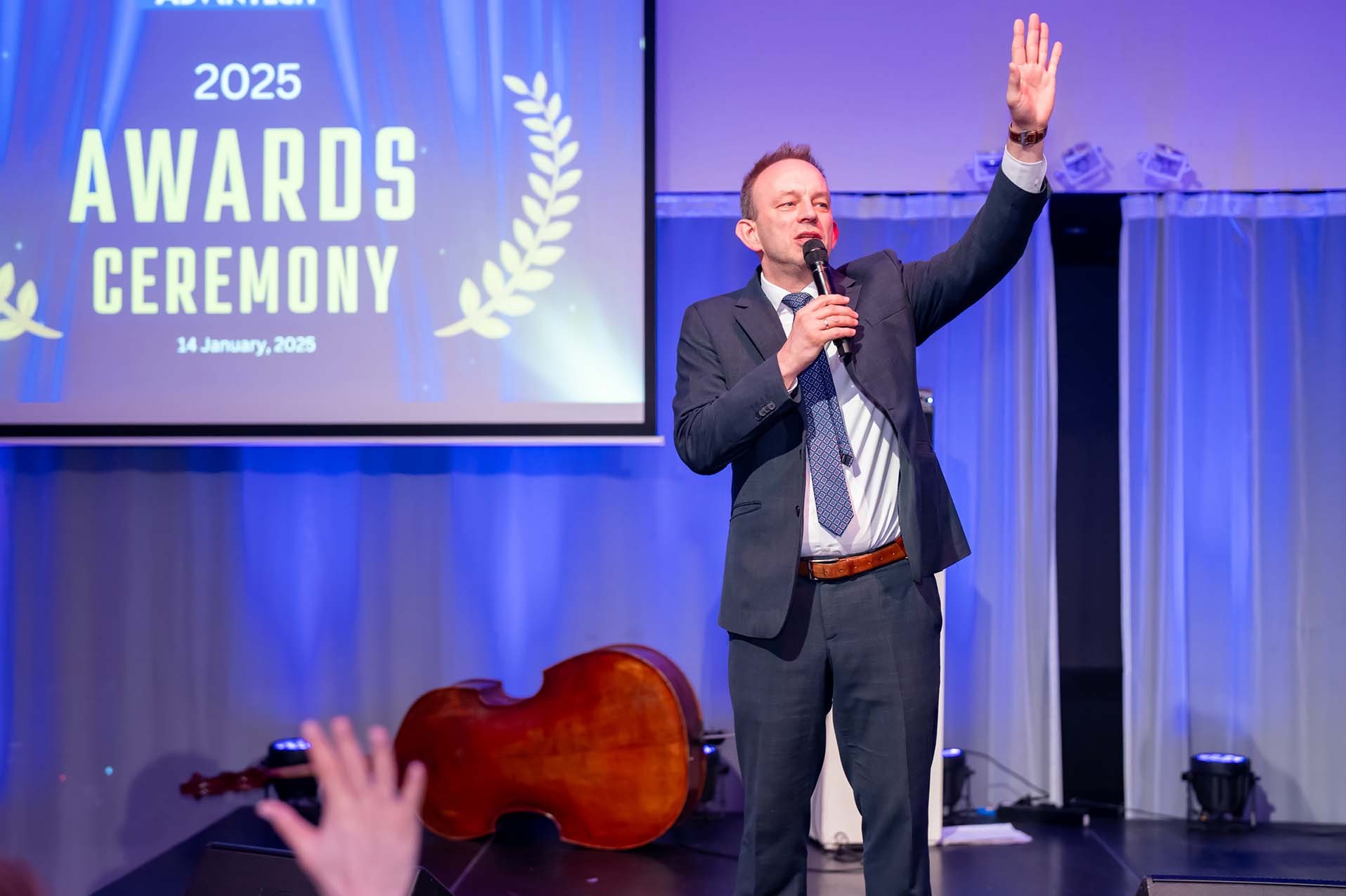 Prezenter w garniturze przemawia na scenie podczas ceremonii wręczenia nagród 2025, z kontrabasem leżącym obok niego.