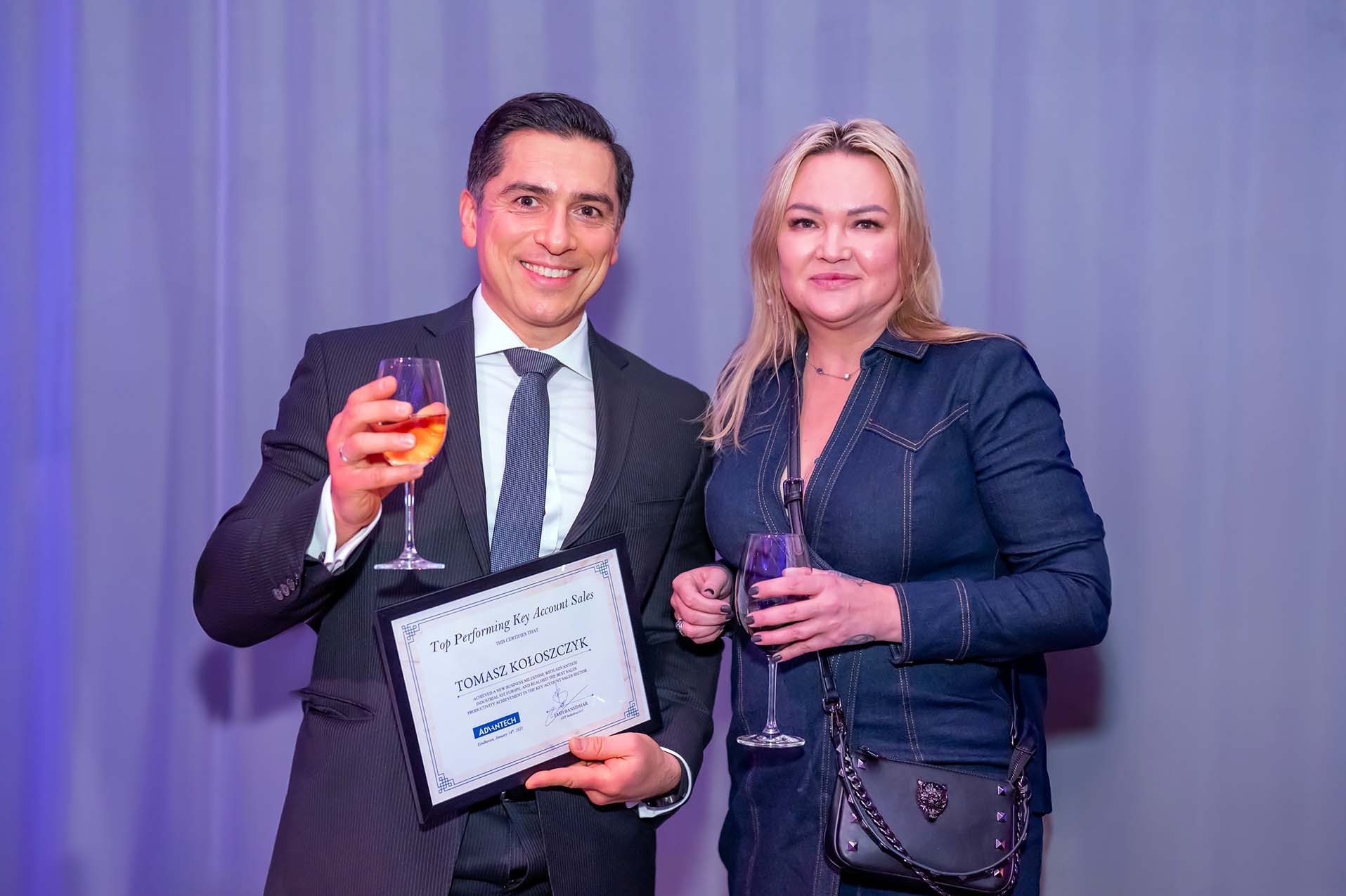 Mężczyzna w garniturze i kobieta w dżinsowej sukience pozują z kieliszkami wina i certyfikatem 'Top Performing Key Account Sales' dla Tomasza Kołoszyka od firmy Advantech.