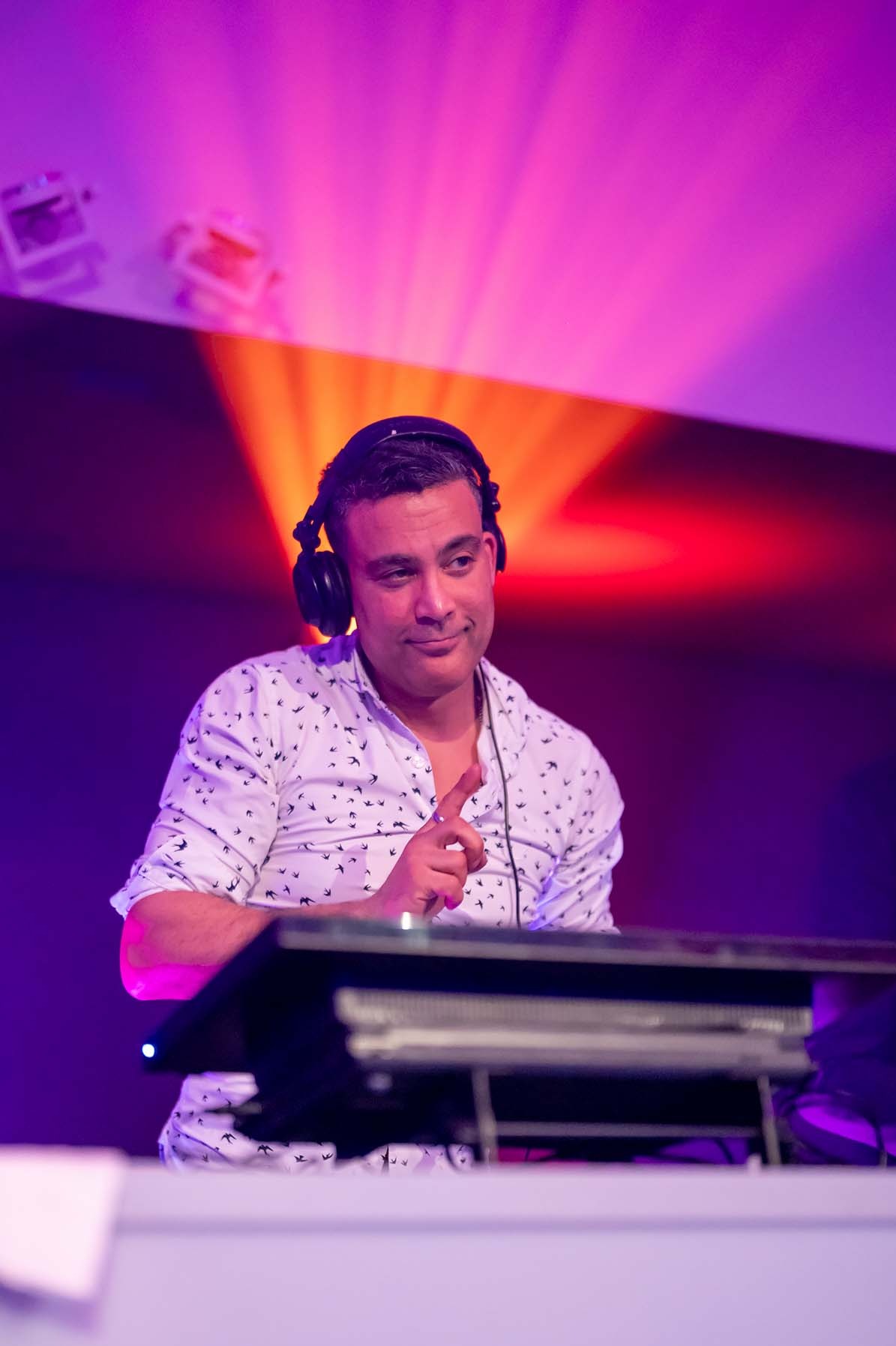 Uśmiechnięty DJ w słuchawkach, w koszuli w ptaszki, z uniesionym palcem, za konsoletą, w fioletowo-pomarańczowym oświetleniu.