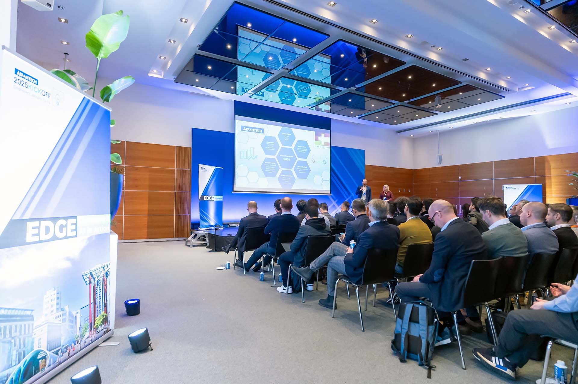 Sala konferencyjna z widokiem na prelegenta i publiczność podczas prezentacji Advantech 2025 Kickoff, z ekranem wyświetlającym dane i logo, oraz roll-upy reklamowe po bokach.