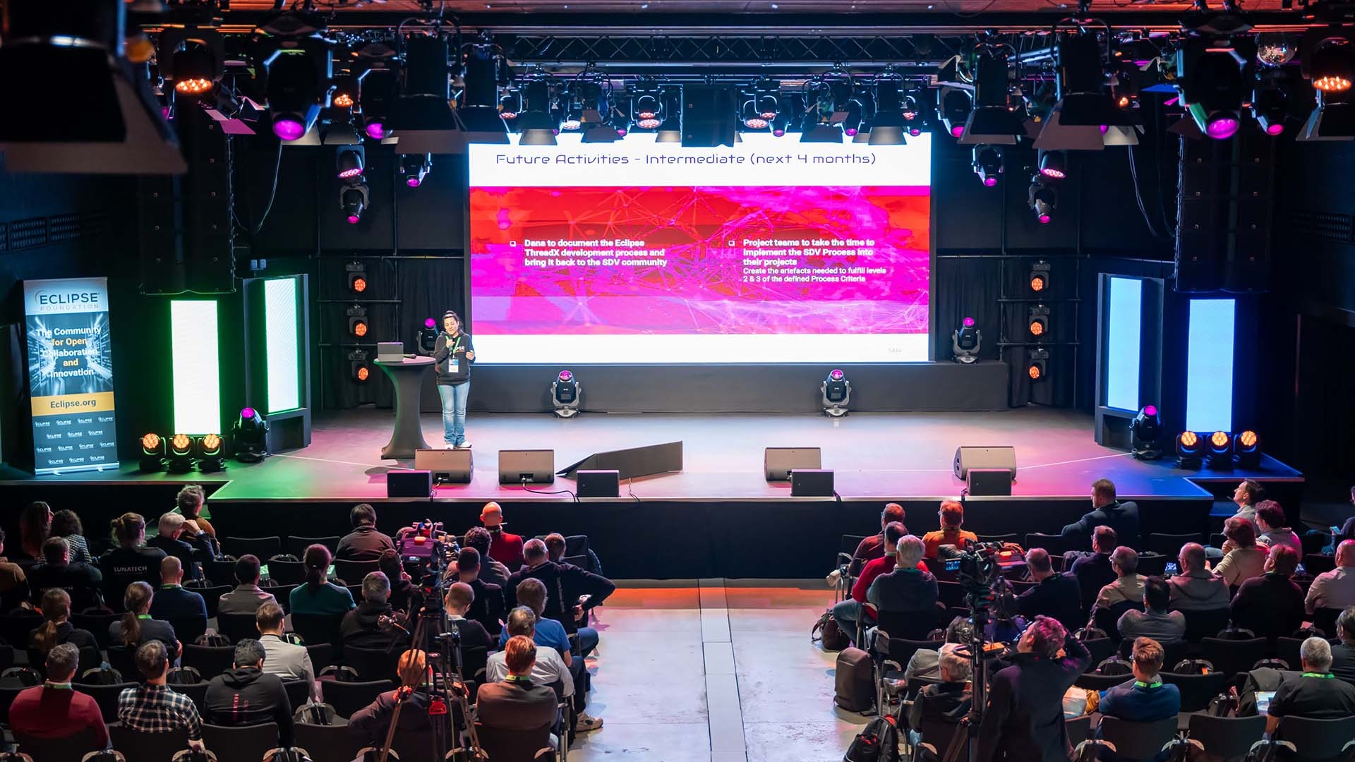 Prelegentka prezentuje slajd 'Future Activities' na scenie podczas konferencji, widownia słucha, oświetlenie sceniczne i baner Eclipse Foundation po lewej stronie.