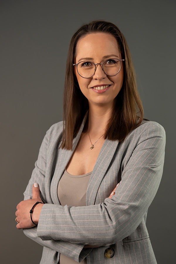 Portret uśmiechniętej kobiety w okularach i szarym garniturze w prążki, z rękami skrzyżowanymi na piersiach, na jednolitym szarym tle.