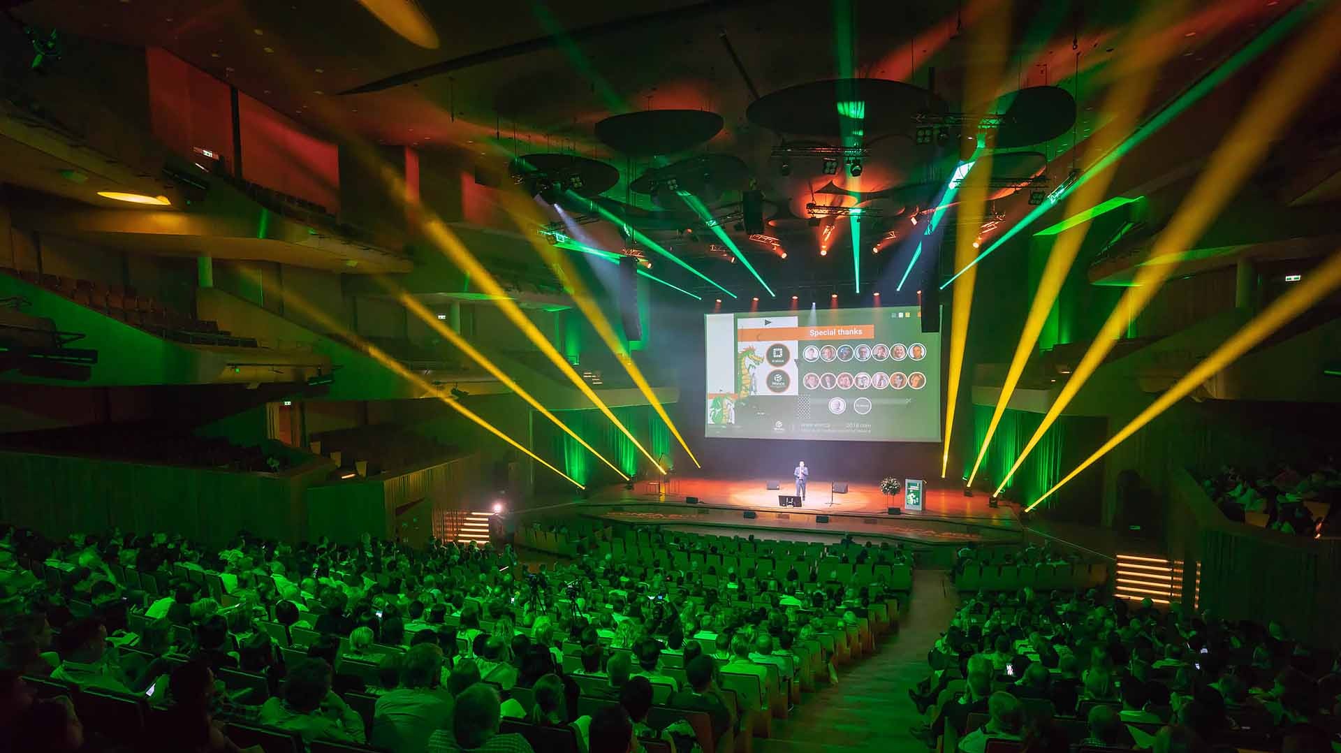 Sala konferencyjna wypełniona publicznością, oświetlona zielonym i żółtym światłem, z prelegentem na scenie i ekranem wyświetlającym prezentację z logotypami i zdjęciami osób.