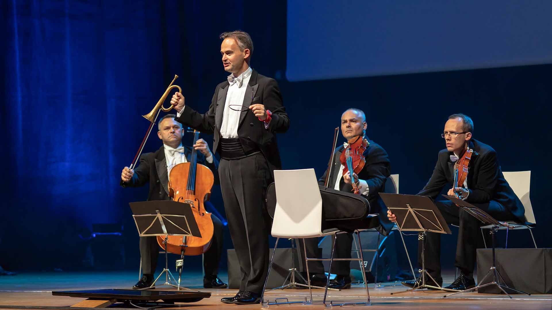 Orkiestra symfoniczna podczas występu: dyrygent w smokingu z batutą i okularami w ręku, trębacz z trąbką sygnałową, wiolonczelista i dwaj skrzypkowie na scenie.