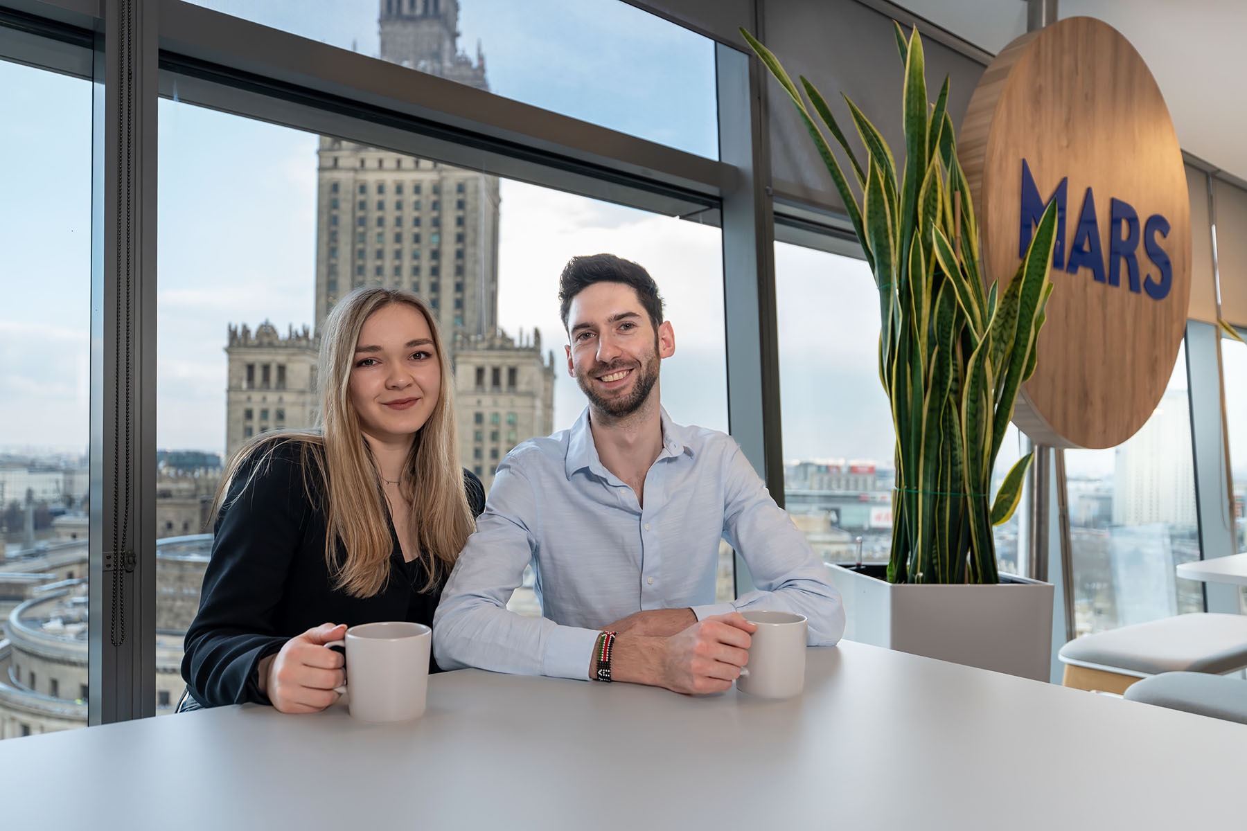 Dwoje uśmiechniętych pracowników biurowych siedzi przy stole z kubkami, z widokiem na Pałac Kultury i Nauki w Warszawie, obok logo firmy MARS i doniczki z rośliną.