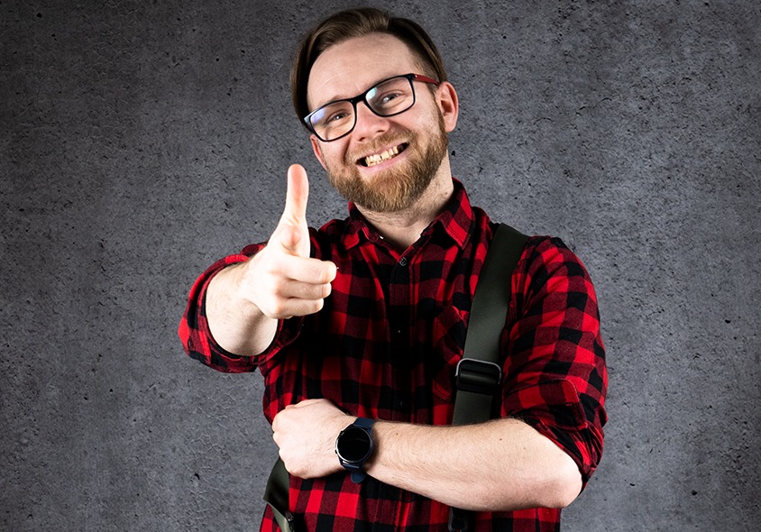 Uśmiechnięty mężczyzna w okularach i czerwono-czarnej koszuli w kratę, z plecakiem na ramieniu, pokazuje kciuk w górę na tle szarej ściany.