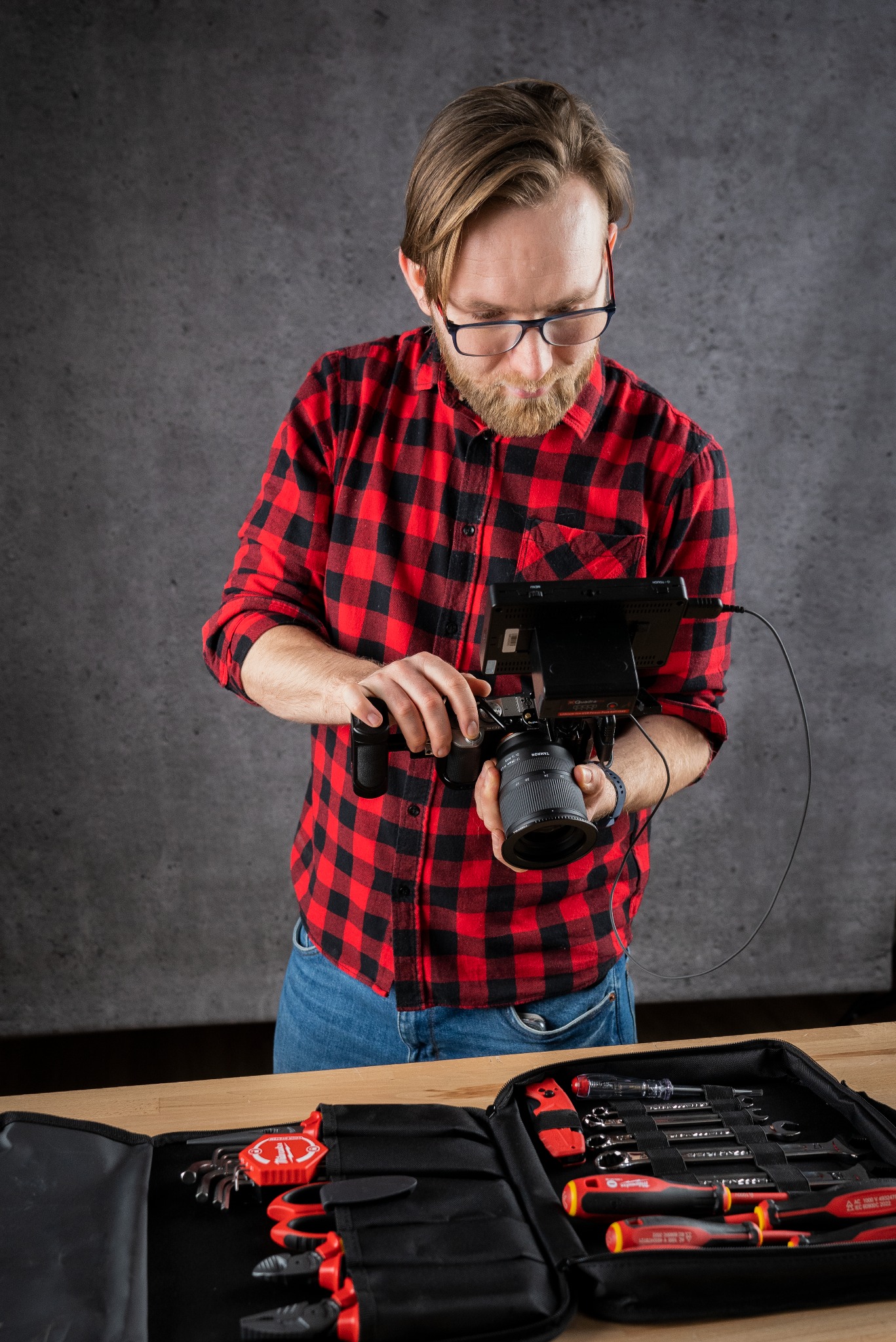 Mężczyzna z brodą w okularach i czerwonej koszuli w kratę trzyma kamerę z podłączonym monitorem, otoczony narzędziami w czarnym organizerze.