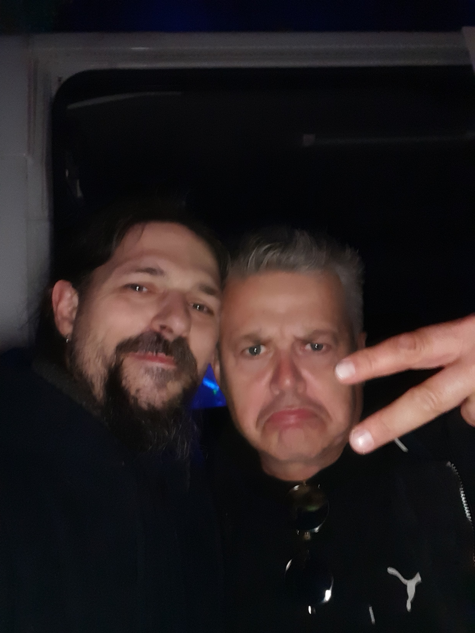 Portret dwóch mężczyzn w słabym oświetleniu, jeden z brodą i kolczykiem, drugi z gestem V, obaj w ciemnych ubraniach