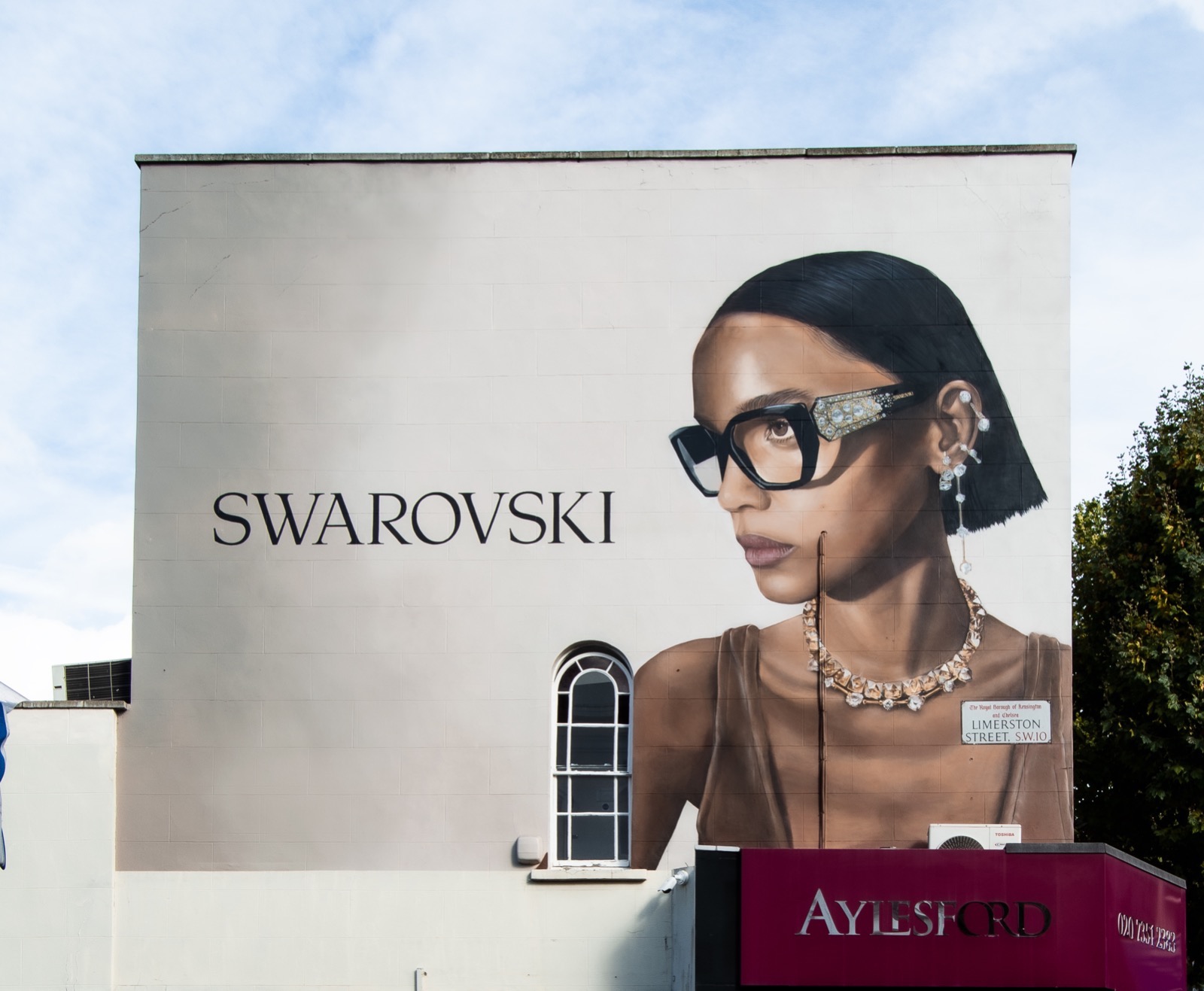 Reklama na ścianie budynku przedstawia portret kobiety w okularach i biżuterii Swarovski, obok widnieje logo firmy.