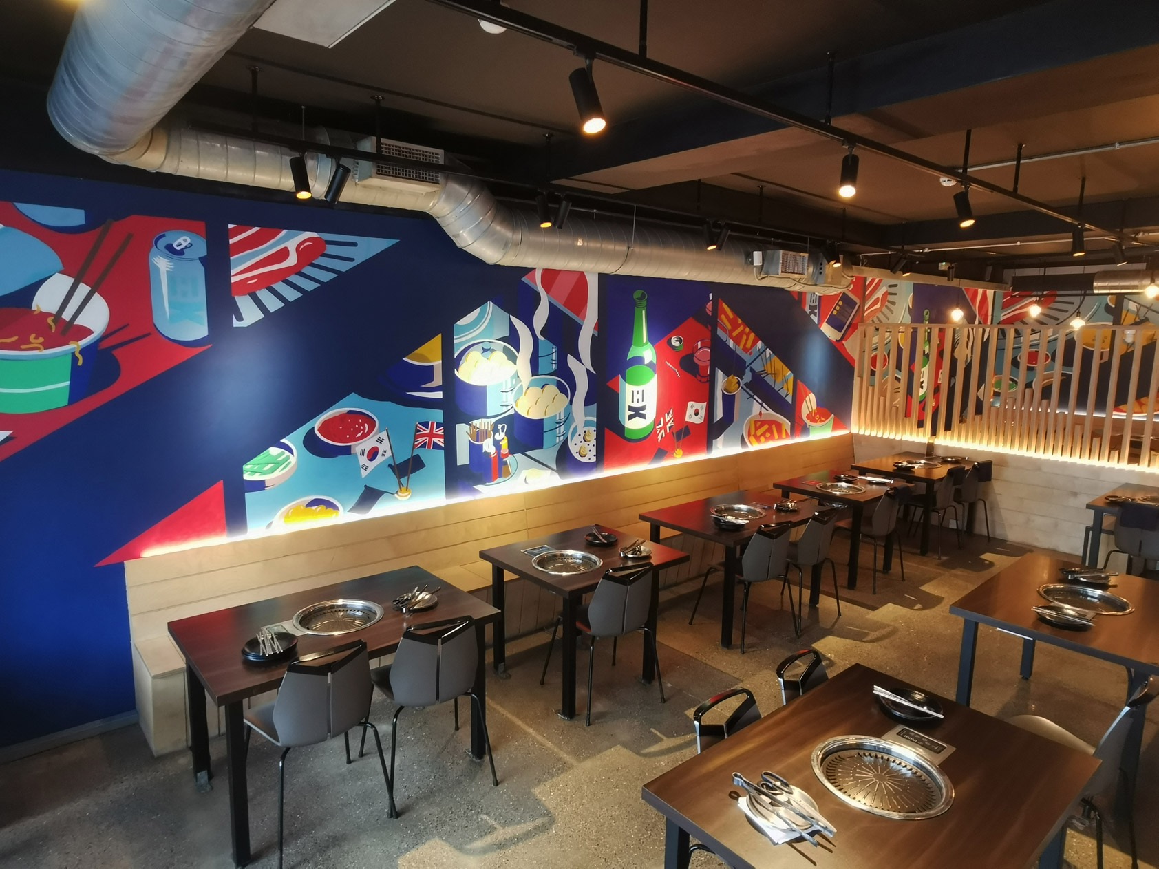 Wnętrze restauracji z muralem przedstawiającym potrawy i flagi różnych krajów, ciemne stoły z metalowymi grillami, szare krzesła, oświetlenie punktowe i drewniane panele.