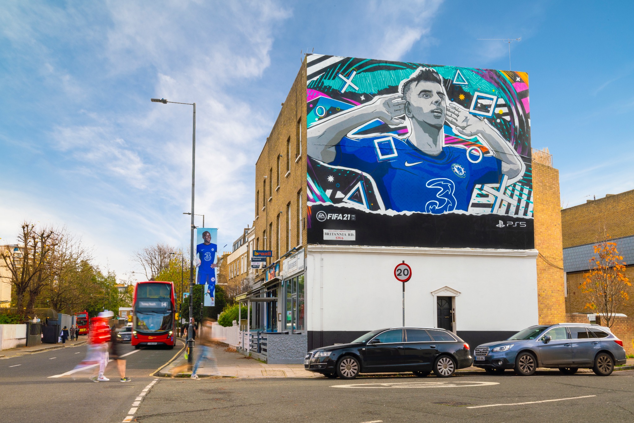 Wielkoformatowa reklama piłkarza w barwach Chelsea FC na ścianie budynku w miejskiej scenerii, z widocznymi samochodami i przechodniami.